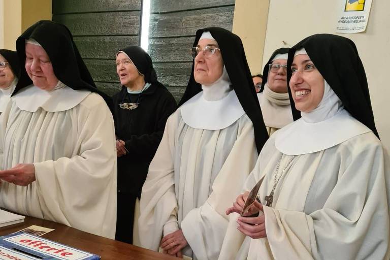 A imagem mostra um grupo de freiras em um evento. Elas estão vestidas com hábitos religiosos, predominantemente brancos e pretos. Algumas freiras estão sorrindo e outras parecem estar em um momento de reflexão. Ao fundo, é possível ver mais freiras e uma mesa com documentos e materiais relacionados ao evento.