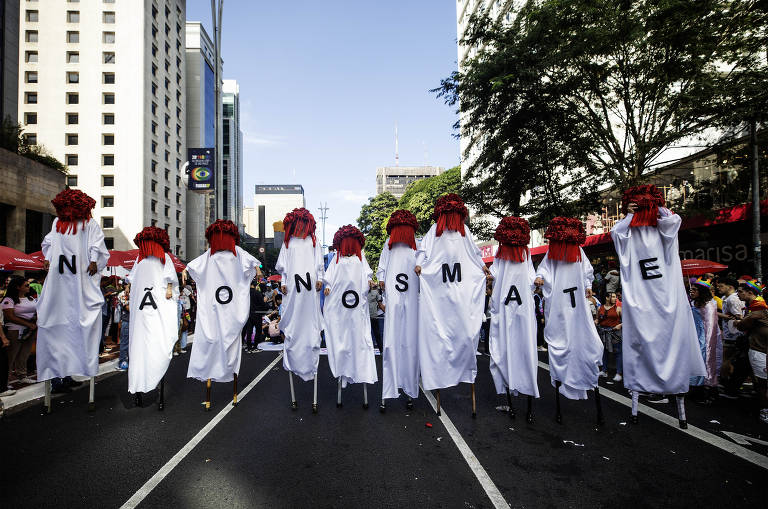 A imagem mostra um grupo de pessoas vestidas com roupas brancas e cabeças cobertas por lenços vermelhos, cada um tem na roupa uma letra. Juntas, elas formam a frase 'NÃO NOS MATE'. Eles estão em uma rua, com prédios altos ao fundo e árvores nas laterais. A cena parece ser de uma manifestação ou desfile.