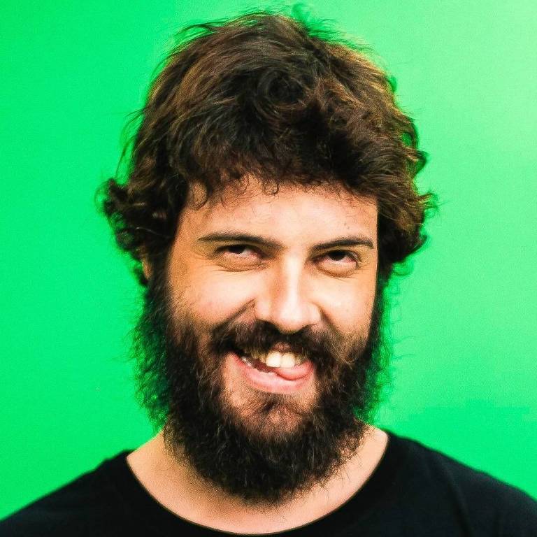 Um homem com cabelo castanho e barba espessa, sorrindo e mostrando a língua. Ele está usando uma camiseta preta e o fundo da imagem é verde.