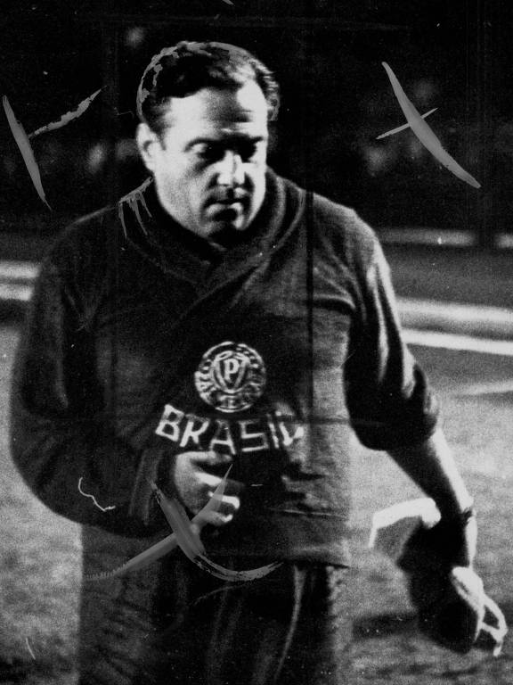 A imagem mostra um jogador de futebol em preto e branco, vestindo um uniforme escuro com um emblema circular no peito que contém as letras 'P' e 'B'. Ele parece estar saindo de campo, com uma expressão séria. O fundo é desfocado, sugerindo um ambiente de jogo. A imagem apresenta marcas de desgaste e arranhões.