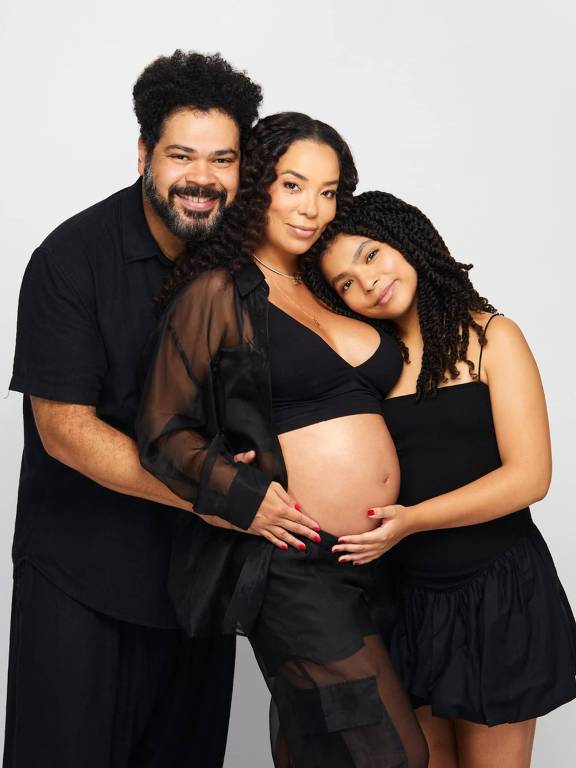 Uma família posando em um estúdio fotográfico. A mulher grávida está no centro, vestindo uma blusa preta e uma saia preta, com uma camisa transparente por cima. Ela está segurando a barriga. À sua esquerda, um homem sorridente, com cabelo crespo e barba, está abraçando-a. À direita, uma jovem com cabelo trançado está encostada na mulher, sorrindo. Todos estão com roupas escuras e o fundo é branco.