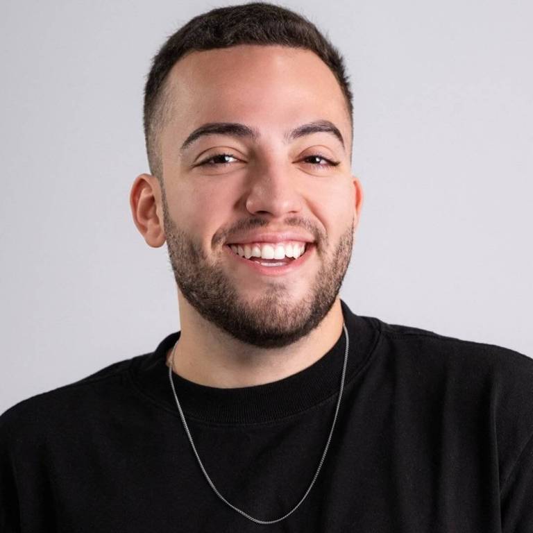 Um homem jovem com cabelo curto e barba, vestindo uma camiseta preta. Ele está sorrindo e olhando para a câmera, com um colar prateado visível em seu pescoço. O fundo é claro e neutro, destacando seu rosto e expressão alegre.