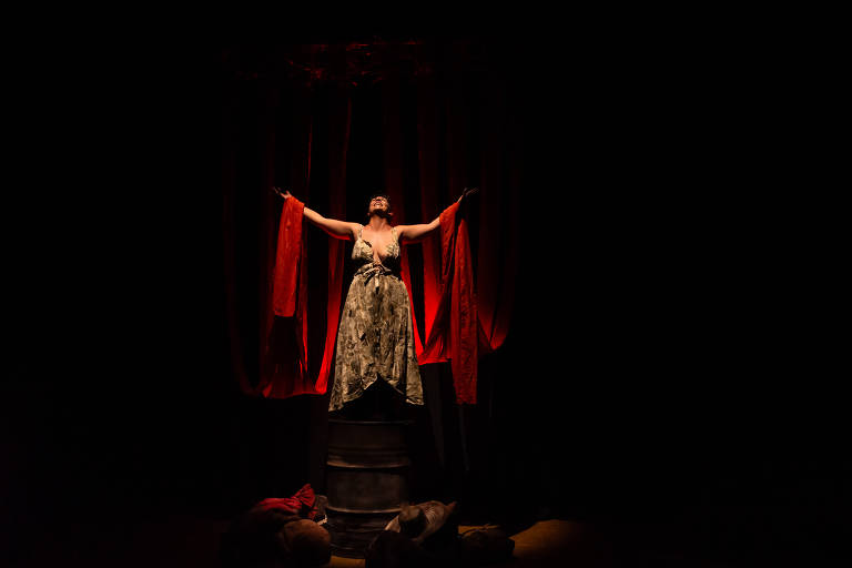 A imagem mostra uma mulher em uma performance teatral, em pé sobre um barril. Ela está vestida com um vestido longo e solto, com um padrão camuflado. A mulher tem os braços abertos, segurando faixas vermelhas que caem ao seu redor. O fundo é escuro, com algumas luzes vermelhas iluminando a cena, criando um contraste dramático. No chão, há objetos que parecem ser roupas ou tecidos amontoados.