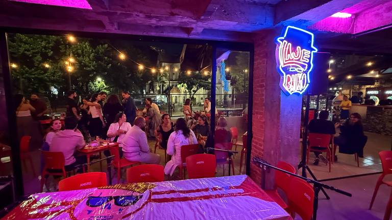 A imagem mostra o interior de um bar com mesas e cadeiras vermelhas. Ao fundo, há um grupo de pessoas conversando e se divertindo. A iluminação é suave, com luzes penduradas e um letreiro de neon azul que diz 'Inje'. A cena é animada, com um ambiente descontraído e acolhedor.