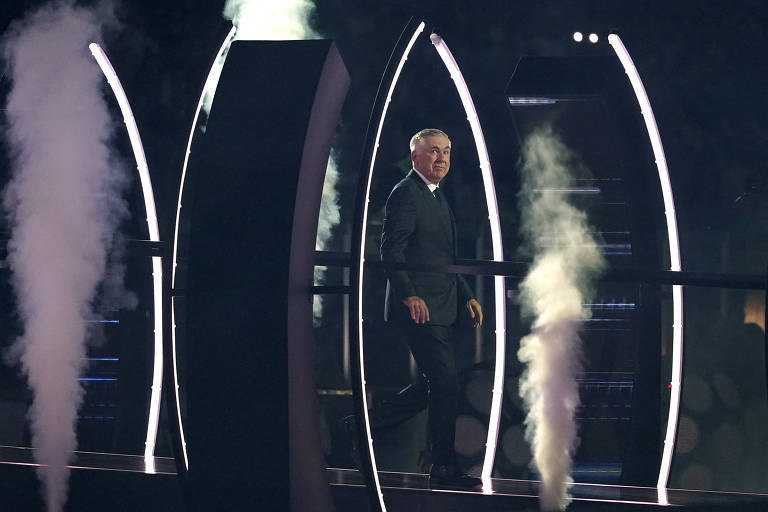 Um homem vestido com um terno escuro caminha por um palco iluminado, cercado por estruturas verticais com luzes brilhantes. Há fumaça saindo de alguns pontos no palco, criando um efeito dramático. O ambiente parece ser de um evento ao vivo, possivelmente uma premiação ou show.