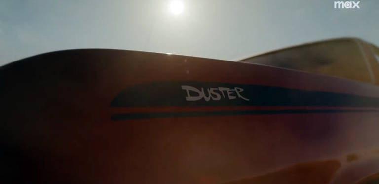 A imagem mostra um close do lado de um carro, onde é visível a inscrição "DUSTER" em uma cor clara, acompanhada de uma faixa azul. O carro é de cor laranja e está sob uma luz solar intensa, com um fundo desfocado que sugere um ambiente ao ar livre.