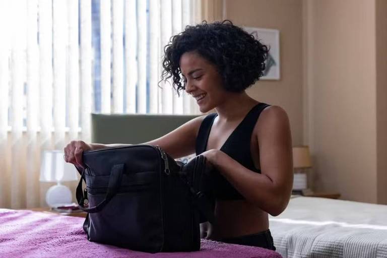 Uma mulher com cabelo cacheado e curto está sorrindo enquanto organiza uma bolsa preta em cima de uma cama. Ela está vestindo um top preto e parece estar em um ambiente interno, com luz natural entrando pelas janelas. Ao fundo, há uma mesa de cabeceira com um abajur e uma parede com uma obra de arte.