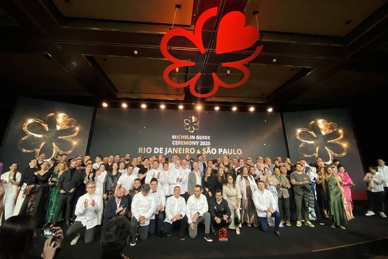 A imagem mostra um grande grupo de pessoas reunidas em um evento, posando para a foto em um palco. Ao fundo, há um grande painel com a inscrição 'MICHELIN GUIDO CEREMONY 2023 RIO DE JANEIRO & SÃO PAULO'. O ambiente é decorado com um grande símbolo de flor e um coração vermelho. As pessoas estão vestidas de maneira formal, algumas em trajes brancos, e outras em roupas coloridas. O público parece estar celebrando um evento especial.