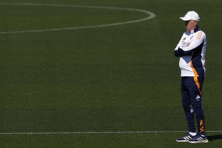 Um homem está em pé em um campo de futebol, com os braços cruzados. Ele usa um uniforme esportivo branco e azul, com detalhes em laranja, e um boné branco. O campo é verde e bem cuidado, com linhas brancas marcando o campo.