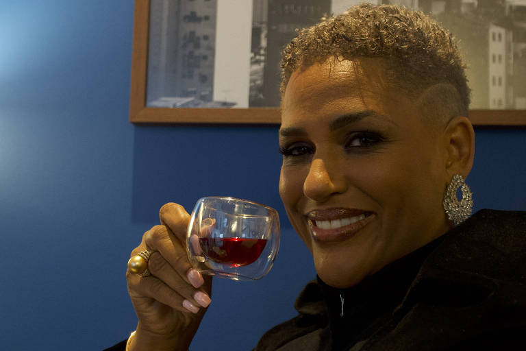 A imagem mostra uma mulher sorridente segurando um pequeno copo com uma bebida vermelha. Ela está usando um casaco preto e tem cabelo curto e grisalho. Ao fundo, há uma parede azul e uma fotografia em preto e branco de uma cidade. A mulher parece estar em um ambiente interno, possivelmente um café ou lounge.