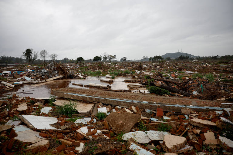 A imagem mostra uma área devastada, com destroços de madeira e telhas espalhados pelo chão. Há uma grande quantidade de entulho e restos de construções, com algumas áreas alagadas. O céu está nublado, sugerindo um clima sombrio, e ao fundo é possível ver uma colina e algumas árvores.