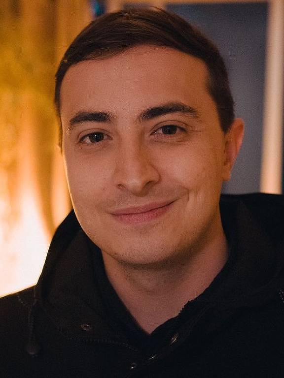 Um jovem homem está sorrindo para a câmera. Ele tem cabelo curto e usa um casaco escuro com capuz. O fundo é iluminado suavemente, com uma luz amarelada e algumas plantas visíveis. A expressão do homem é amigável e relaxada.