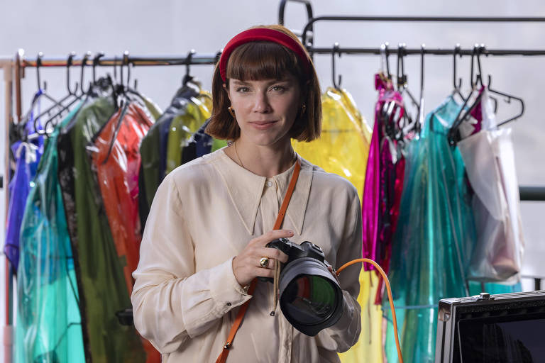 Uma mulher com cabelo curto e castanho, usando uma blusa clara e uma faixa vermelha na cabeça, segura uma câmera fotográfica. Ao fundo, há um cabideiro com várias roupas coloridas, incluindo peças transparentes em tons de verde, vermelho, azul e amarelo. A mulher parece estar em um estúdio de fotografia.