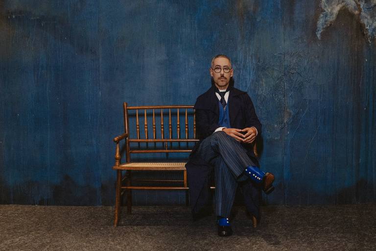 Um homem está sentado em um banco de madeira em um ambiente com uma parede azul ao fundo. Ele usa um terno escuro com uma gravata e meias azuis. O homem tem óculos e uma expressão séria, com as mãos apoiadas sobre os joelhos.