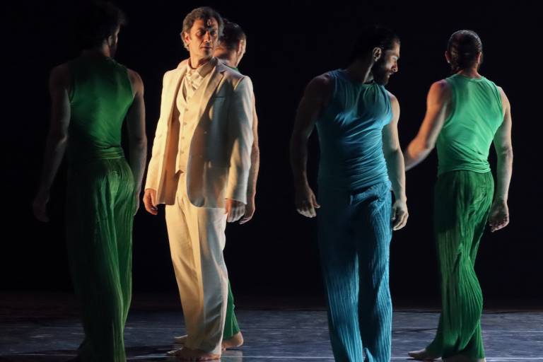 A imagem mostra cinco dançarinos em um palco. Quatro deles estão usando roupas de cores diferentes: dois em verde e dois em azul, enquanto o quinto dançarino, que se destaca, está vestido com um traje claro, composto por um paletó e calças. Os dançarinos estão em várias posições, com alguns de costas e outros de frente, criando uma composição dinâmica. O fundo é escuro, destacando as figuras dos dançarinos.