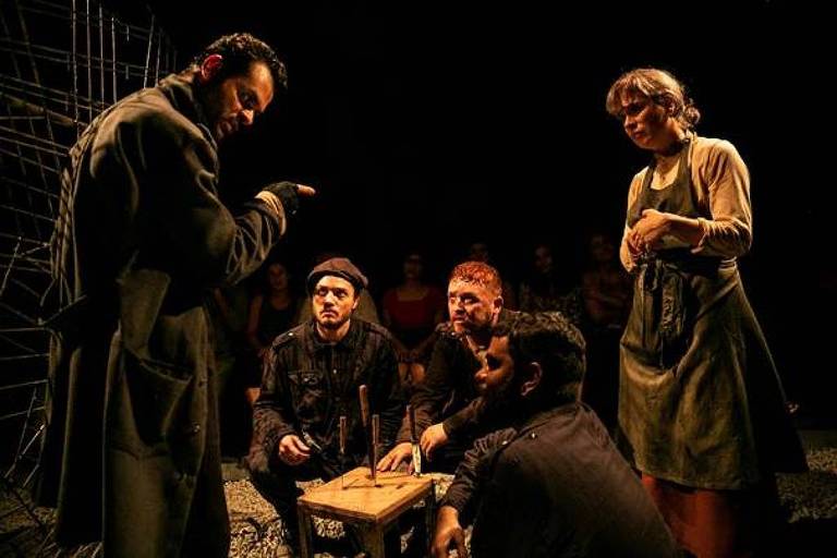 A imagem mostra uma cena de teatro com cinco atores em um ambiente escuro. Um homem em um casaco longo está de pé, gesticulando com a mão, enquanto os outros quatro atores estão sentados ao redor de uma mesa pequena, observando atentamente. Entre eles, há um homem com um chapéu, um com cabelo ruivo e outro com cabelo escuro. Uma mulher está de pé, com as mãos cruzadas, olhando para o grupo. Ao fundo, há uma plateia visível, com algumas pessoas assistindo à cena.