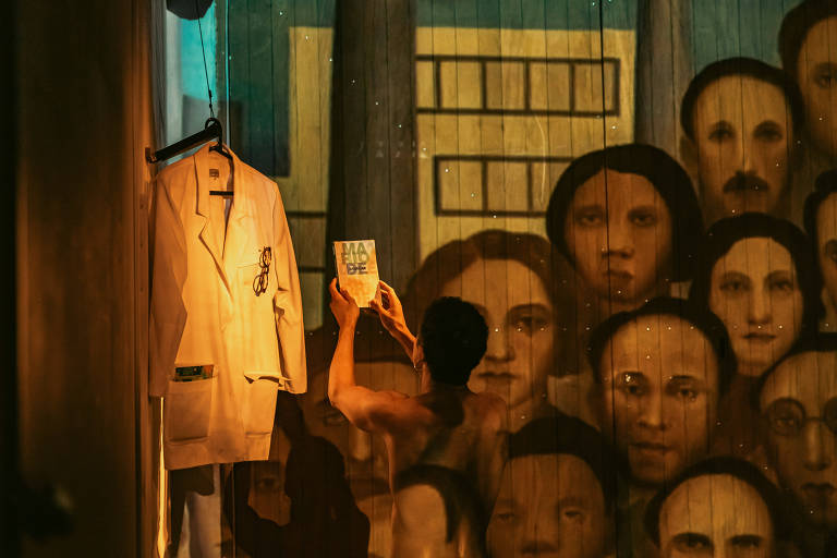 A imagem mostra uma cena com um fundo de pintura que retrata rostos humanos. À esquerda, há um jaleco branco pendurado em um cabide. À direita, uma pessoa está de costas, segurando um livro em direção à pintura. A iluminação é suave, criando um ambiente dramático.