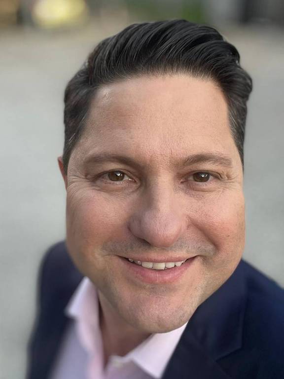 Um homem sorridente está em primeiro plano, com cabelo escuro e bem penteado. Ele usa um paletó escuro e uma camisa clara. O fundo é desfocado, com uma superfície de pedras claras.