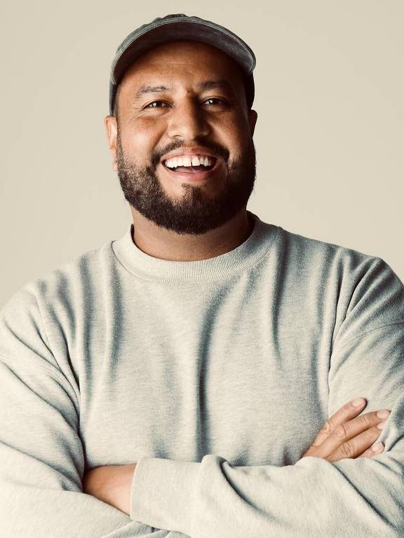 Um homem sorridente está posando com os braços cruzados. Ele usa um suéter cinza e um boné. O fundo é claro e neutro, destacando sua expressão amigável.