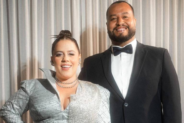 Um casal posando para a foto em um evento formal. A mulher está vestindo um traje prateado brilhante com detalhes texturizados e um decote, enquanto o homem está usando um terno preto com uma gravata borboleta. Ambos estão sorrindo e a mulher está com a mão na cintura. O fundo é uma cortina clara.
