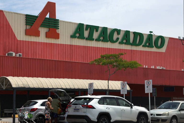 A imagem mostra a fachada de uma loja Atacadão, com um grande letreiro em verde e vermelho. Na frente da loja, há vários carros estacionados, incluindo um carro branco e um SUV. Uma pessoa está ao lado de um dos carros, aparentemente organizando as compras. O céu está claro e a iluminação é de dia.