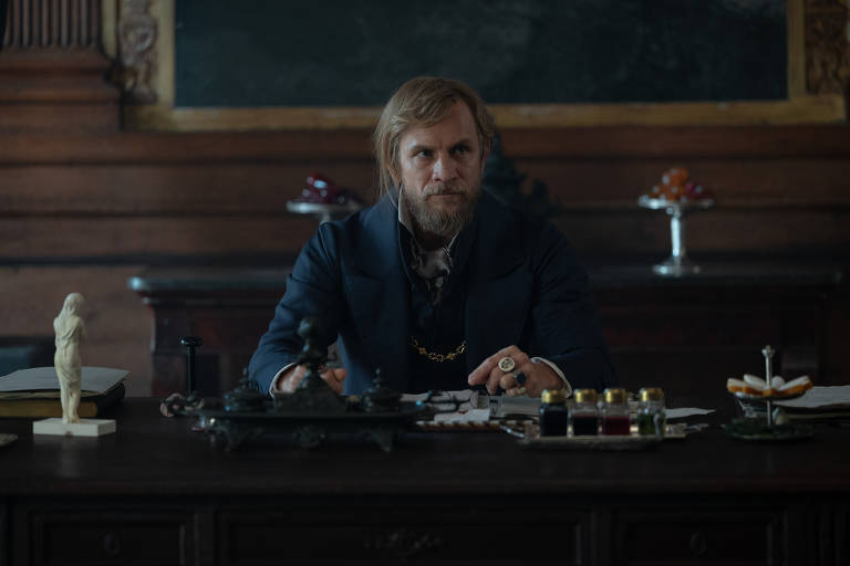 A imagem mostra um homem sentado em uma mesa de escritório, vestindo um traje vitoriano. Ele tem cabelo loiro e barba, e está olhando para a frente com uma expressão séria. Na mesa, há vários objetos, incluindo um livro, uma estatueta, um suporte com frascos coloridos e um prato com doces. O ambiente é decorado com madeira escura e um fundo que sugere um espaço de trabalho antigo.