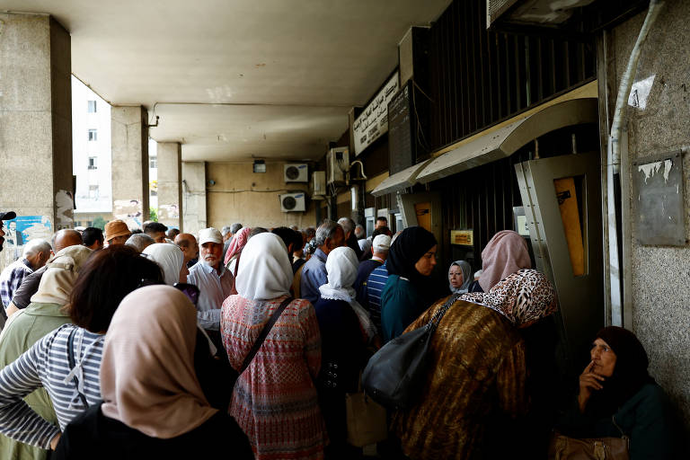 A imagem mostra uma grande fila de pessoas em frente a um caixa eletrônico. A maioria das pessoas está usando roupas tradicionais e coberturas de cabeça. O ambiente parece ser urbano, com paredes de concreto e alguns aparelhos de ar-condicionado visíveis. A fila é composta principalmente por adultos e idosos, demonstrando uma cena de espera em um local público.