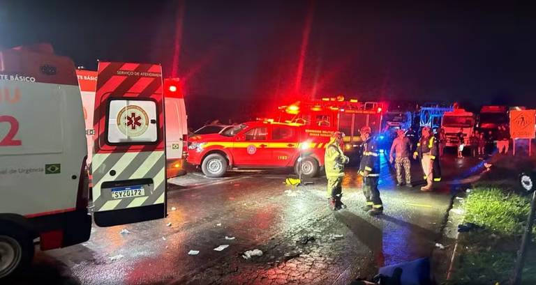 A imagem mostra uma cena de acidente à noite, com várias ambulâncias e veículos de emergência. Há luzes vermelhas piscando e pessoas em trajes de emergência, como bombeiros e paramédicos, trabalhando na área. O chão está molhado, possivelmente devido à chuva, e há alguns detritos visíveis na estrada. Ao fundo, é possível ver caminhões estacionados e um sinal de trânsito.