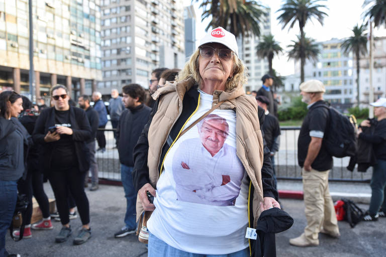 Uma mulher idosa está em um protesto, vestindo uma camiseta branca com a imagem de um homem sorridente. Ela usa um colete bege e um boné branco com a palavra 'Gol'. Ao fundo, há um grupo de pessoas e edifícios altos, além de palmeiras.
