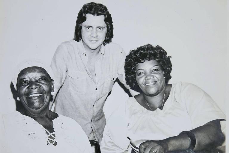 A imagem em preto e branco mostra três pessoas sorrindo. À esquerda, uma mulher idosa com pele escura e cabelo curto, vestindo uma blusa branca com detalhes. No centro, um homem com cabelo longo e liso, usando uma camisa clara. À direita, uma mulher negra e de cabelo cacheado, vestindo uma blusa branca. O fundo é simples e sem detalhes.