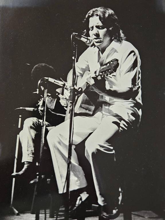 A imagem em preto e branco mostra um artista sentado em um palco, tocando violão e cantando em um microfone. Ele está vestido com uma camisa clara e calças brancas. Ao fundo, um outro músico está visível, sentado e tocando um instrumento. A iluminação do palco destaca os dois artistas, criando um ambiente intimista.