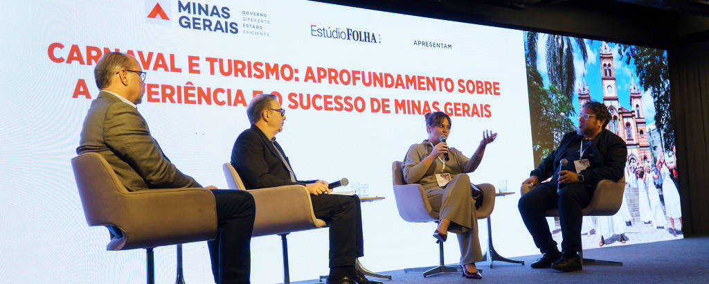 A imagem mostra um painel de debate com três pessoas sentadas em cadeiras em um palco. Ao fundo, há um grande telão com o texto 'CARNAVAL E TURISMO: APROFUNDAMENTO SOBRE A EXPERIÊNCIA E SUCESSO DE MINAS GERAIS' e os logotipos de 'MINAS GERAIS' e 'Estúdio Ponta'. As pessoas no painel estão vestidas de forma formal, com um homem de terno cinza, um homem de terno preto e uma mulher com blusa clara. O ambiente é moderno e bem iluminado.