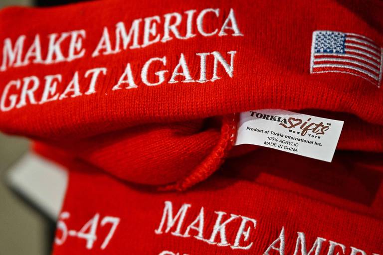 A imagem mostra bonés vermelhos empilhados, com o slogan "MAKE AMERICA GREAT AGAIN" bordado em branco. Um dos bonés exibe uma pequena bandeira dos Estados Unidos. Também é visível uma etiqueta que diz "Torkita Gifts" e informações sobre a fabricação na China.