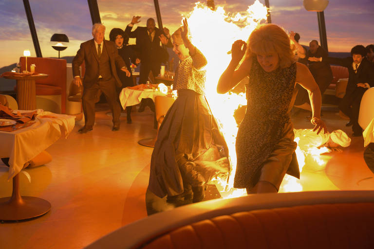 A imagem mostra uma cena caótica em um restaurante, onde várias pessoas estão em pânico. Uma mulher está correndo com fogo ao seu redor, enquanto outras pessoas, incluindo um homem de terno e uma mulher com cabelo escuro, parecem estar tentando escapar. O ambiente é iluminado por uma luz laranja, possivelmente do pôr do sol, e as mesas estão decoradas com toalhas e pratos. O chão é de um material claro e liso, e há uma sensação de urgência e desespero na cena.