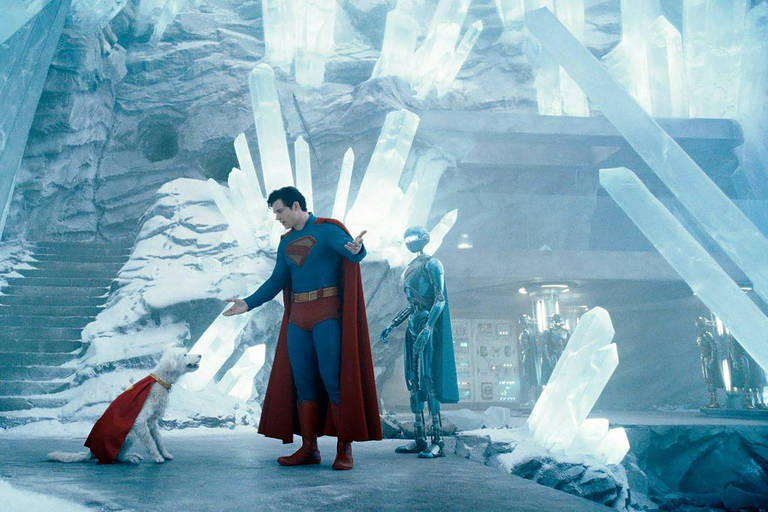 A imagem mostra um personagem vestido como super-herói, com um traje azul e uma capa vermelha, interagindo com um cachorro que também usa uma capa vermelha. Ao fundo, há uma estrutura de cristais de gelo e um robô prateado. O ambiente é gelado e iluminado, com uma atmosfera futurista.