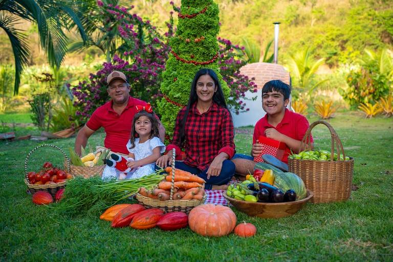 Uma família composta por um homem, uma mulher, uma menina e um menino está sentada em um gramado verde. Eles estão cercados por cestas cheias de frutas e vegetais, incluindo tomates, cenouras, abóbora, bananas e outros produtos. Ao fundo, há uma árvore ornamental e plantas floridas, além de uma estrutura branca que parece ser uma churrasqueira. Todos estão sorrindo e vestindo roupas casuais, com a mulher usando uma blusa xadrez e os homens camisetas vermelhas.