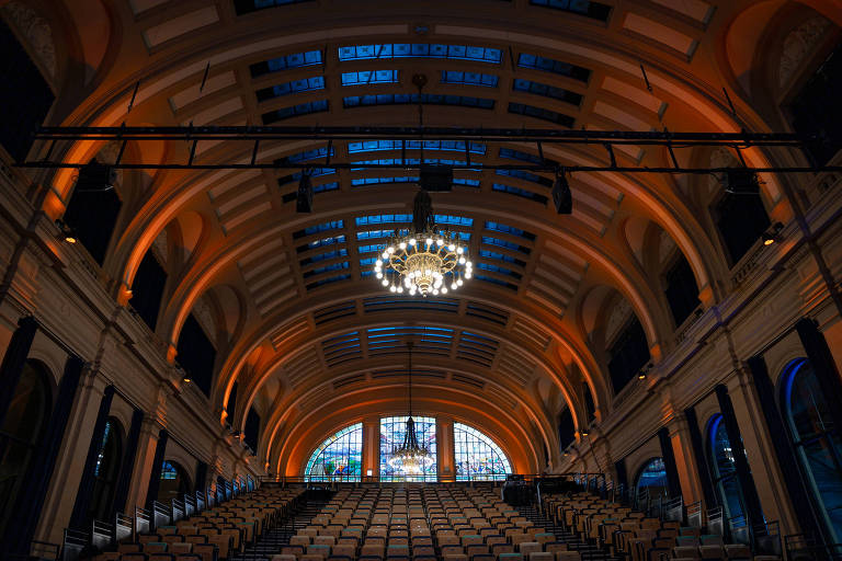 A imagem mostra o interior de um auditório com um teto alto e abobadado. O espaço é iluminado por luzes laranjas e possui uma grande candelabro central. As paredes são decoradas e há uma grande janela ao fundo, com vitrais. As cadeiras estão dispostas em fileiras, voltadas para o palco.