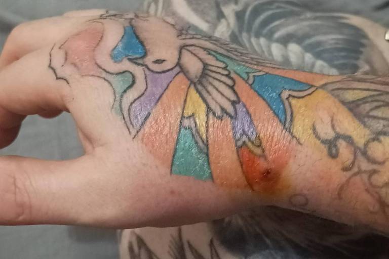 A imagem mostra a mão de uma pessoa com uma tatuagem colorida no dorso. A tatuagem apresenta várias cores, incluindo azul, laranja, verde e rosa, formando um design que parece ter elementos de nuvens e raios. A pele ao redor da tatuagem parece um pouco vermelha, possivelmente indicando que a tatuagem é recente. Ao fundo, é possível ver outros desenhos de tatuagens na parte inferior do braço.