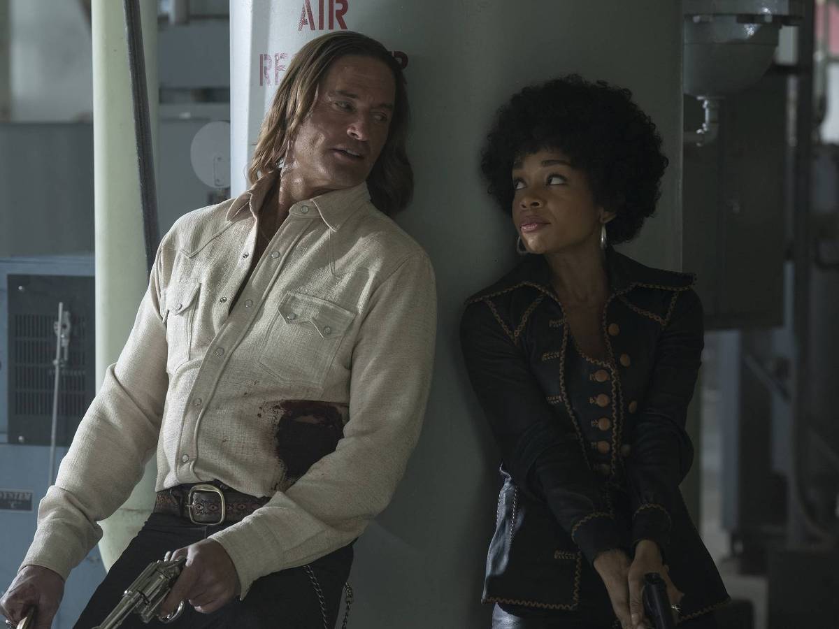 Cena da primeira temporada da série Duster com Josh Holloway e Rachel Hilson *** Local Caption *** A imagem mostra dois personagens em uma cena de ação. O homem à esquerda tem cabelo longo e usa uma camisa clara com manchas de sangue, enquanto a mulher à direita tem cabelo crespo e usa uma jaqueta preta com detalhes em dourado. Ambos estão encostados em uma coluna, segurando armas e olhando um para o outro, em um ambiente industrial.