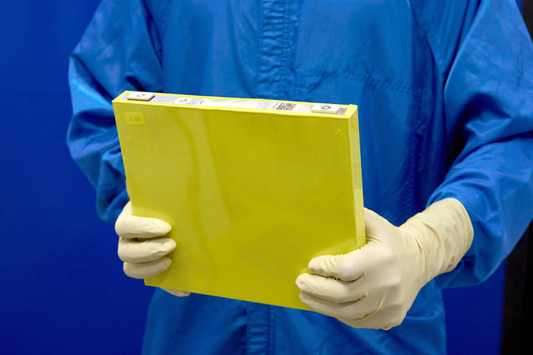 A imagem mostra uma pessoa vestindo um traje azul e luvas brancas, segurando um pacote amarelo. O fundo é azul, sugerindo um ambiente controlado, possivelmente de laboratório. O pacote parece ser de material rígido e possui uma etiqueta visível na parte superior. Trata-se de um módulo de bateria para carro elétrico