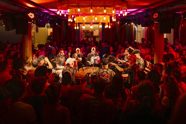 A imagem mostra uma apresentação musical em um ambiente intimista, com um grande público ao redor. No centro, um grupo de músicos toca instrumentos, enquanto a plateia observa atentamente. A iluminação é suave, com luzes vermelhas e pendentes, criando uma atmosfera aconchegante. O público é diversificado e parece estar envolvido na performance.