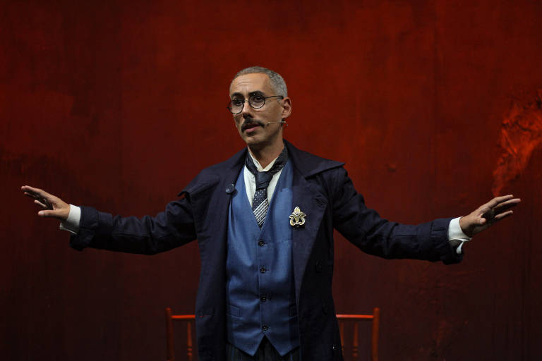 Um homem está em um palco, vestido com um traje formal que inclui um paletó escuro e um colete azul. Ele tem cabelo grisalho e usa óculos. O fundo do palco é de uma cor avermelhada, e ele está com os braços abertos, como se estivesse se dirigindo a uma audiência. Há uma cadeira de madeira ao fundo.