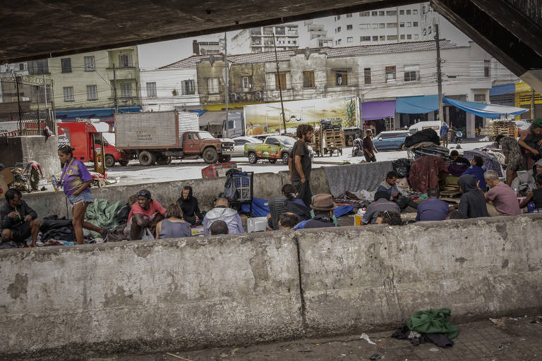 A imagem mostra um grupo de pessoas em uma área urbana, possivelmente sob um viaduto. Algumas pessoas estão sentadas no chão, cercadas por cobertores e pertences pessoais. Ao fundo, há veículos estacionados e edifícios, com uma atmosfera de descaso. A cena retrata a realidade de pessoas em situação de vulnerabilidade social.