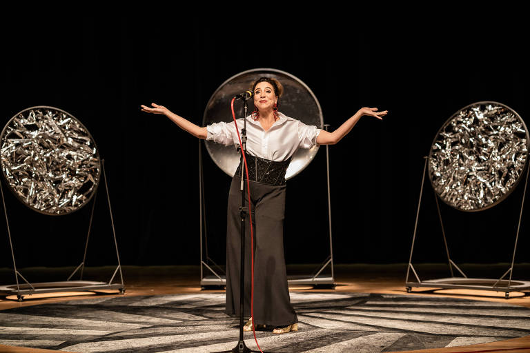 Uma artista se apresenta em um palco, usando uma blusa branca e uma saia longa preta. Ela está em uma pose expressiva, com os braços abertos, em frente a um microfone. Ao fundo, há dois grandes círculos prateados que refletem a luz, e o chão do palco é decorado com um padrão geométrico. O ambiente é escuro, destacando a artista e os elementos ao redor.
