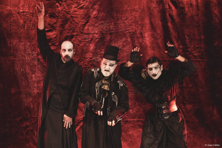 A imagem mostra três artistas de teatro em uma apresentação. Eles estão vestidos com roupas escuras e maquiagem dramática, com rostos pintados em tons brancos e pretos. O fundo é de um tecido vermelho, criando um contraste marcante. Os artistas estão em poses expressivas, com um deles levantando a mão, enquanto os outros têm expressões intensas e gestos teatrais.