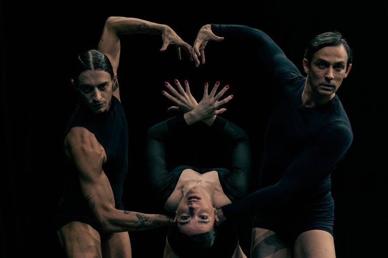 A imagem mostra três dançarinos em uma apresentação de dança contemporânea. Dois homens estão posicionados em pé, um de cada lado, enquanto uma mulher está suspensa entre eles, com a cabeça para baixo. Os dançarinos estão vestidos com roupas justas e escuras, e suas poses expressam movimento e emoção. O fundo é escuro, destacando os corpos e as formas dos dançarinos.