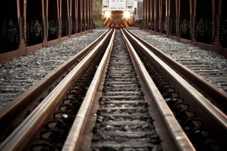 Imagem mostra, em primeiro plano, trilhos ferroviários; ao fundo, parte de uma locomotiva, com os faróis acesos, indica se aproximar