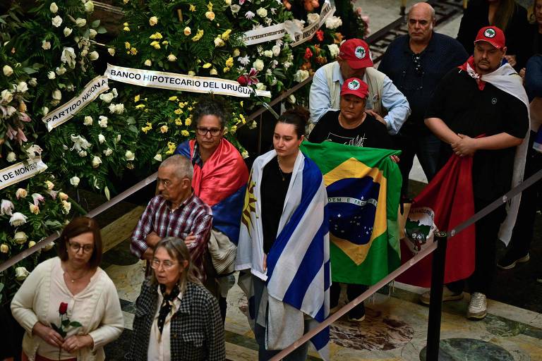 A imagem mostra um grupo de pessoas participando de uma manifestação. Algumas pessoas estão segurando bandeiras do Brasil e do Uruguai, enquanto outras usam faixas e bonés com símbolos de um partido político. O ambiente parece ser um espaço público, decorado com flores. As pessoas estão em fila, algumas com expressões sérias, e há uma mulher na frente segurando uma rosa.