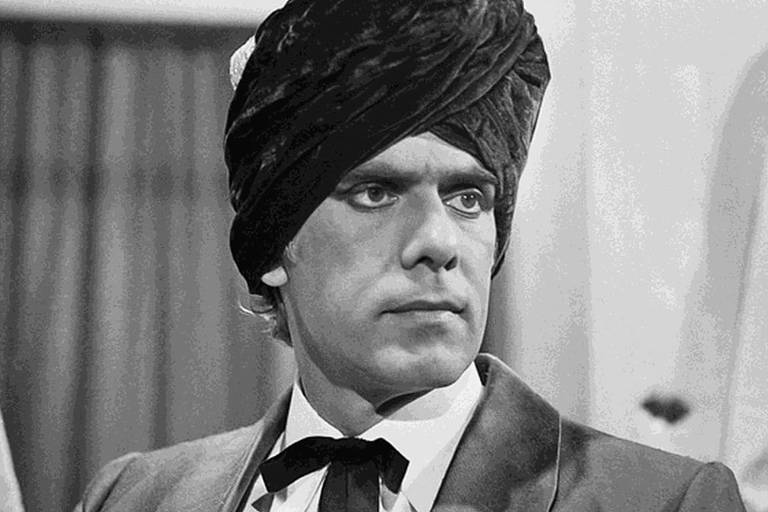 A imagem mostra um homem com um turbante escuro e um terno claro. Ele tem uma expressão séria e está olhando para frente. O fundo é desfocado, com cortinas que sugerem um ambiente interno.