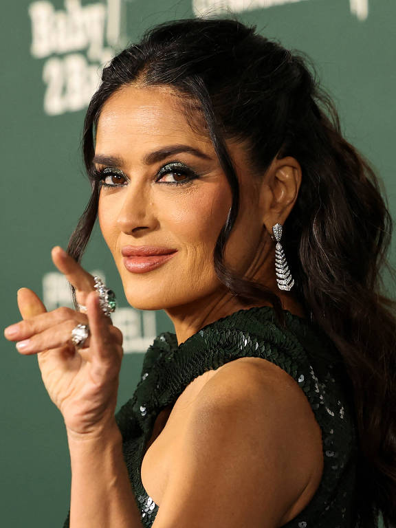 Uma mulher com cabelo longo e solto, usando um vestido verde brilhante, está posando para a câmera. Ela está fazendo um gesto com a mão, levantando o dedo indicador, e sorrindo. O fundo é de cor verde escura com texto em branco que parece ser parte de um evento ou premiere.